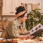 A woman looks at a cookbook while preparing pastry in a cozy kitchen setting with flowers nearby.