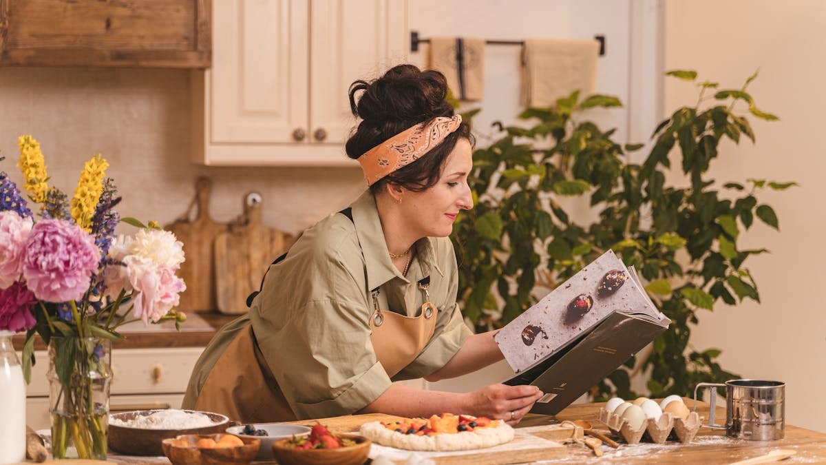 A woman looks at a cookbook while preparing pastry in a cozy kitchen setting with flowers nearby.